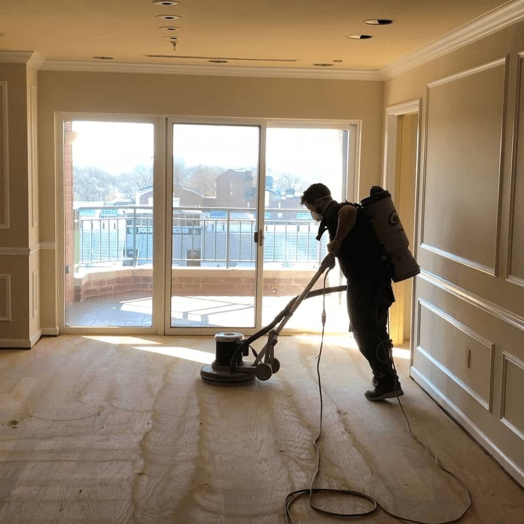 A worker sands a hardwood floor in a room with large windows overlooking a balcony.