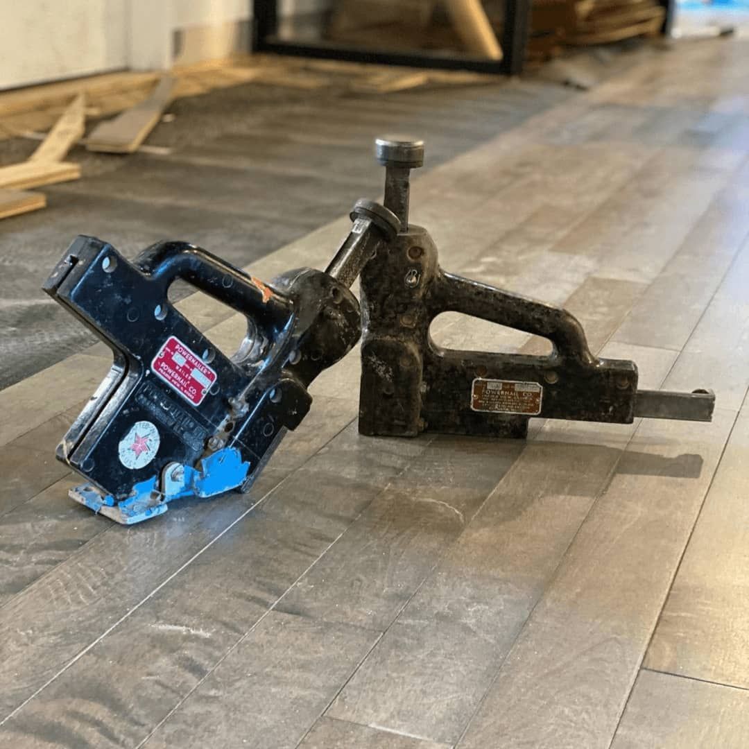 Two floor-installing tools on a gray wood floor: a black tool with a blue edge and another darker tool with a screw.