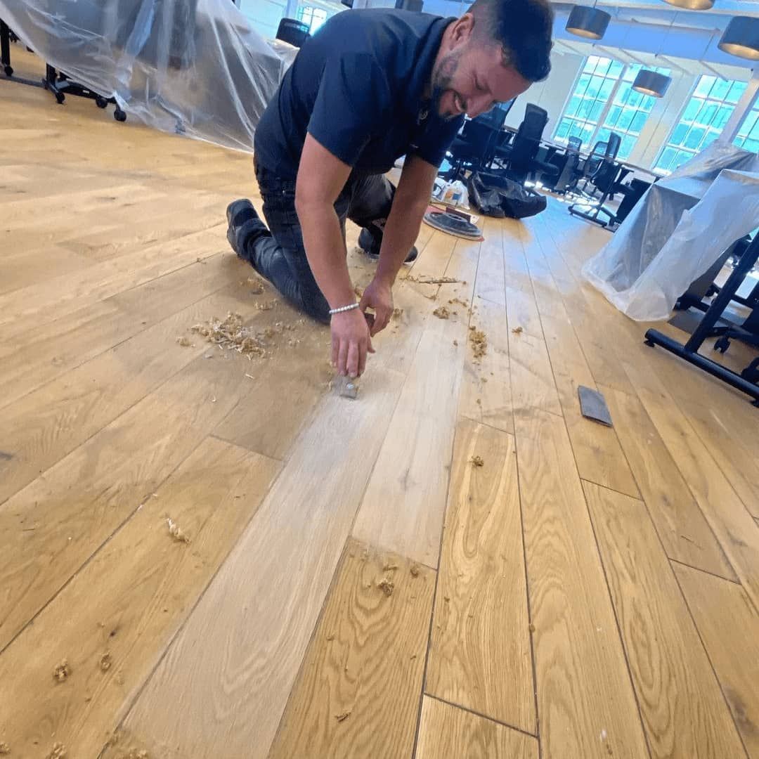 Man kneeling on hardwood floor, using a scraper to remove something. Office setting.
