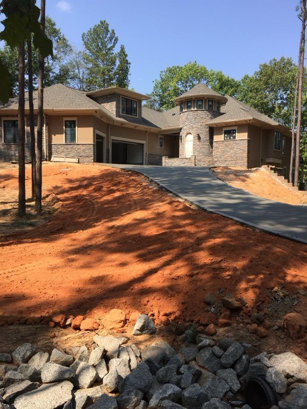A tan, multi-story house with a stone turret and driveway under construction sits on a large, red-dirt slope.