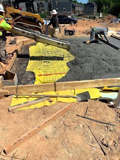 Construction workers pour concrete into a wooden frame over a yellow vapor barrier on a sunny job site.