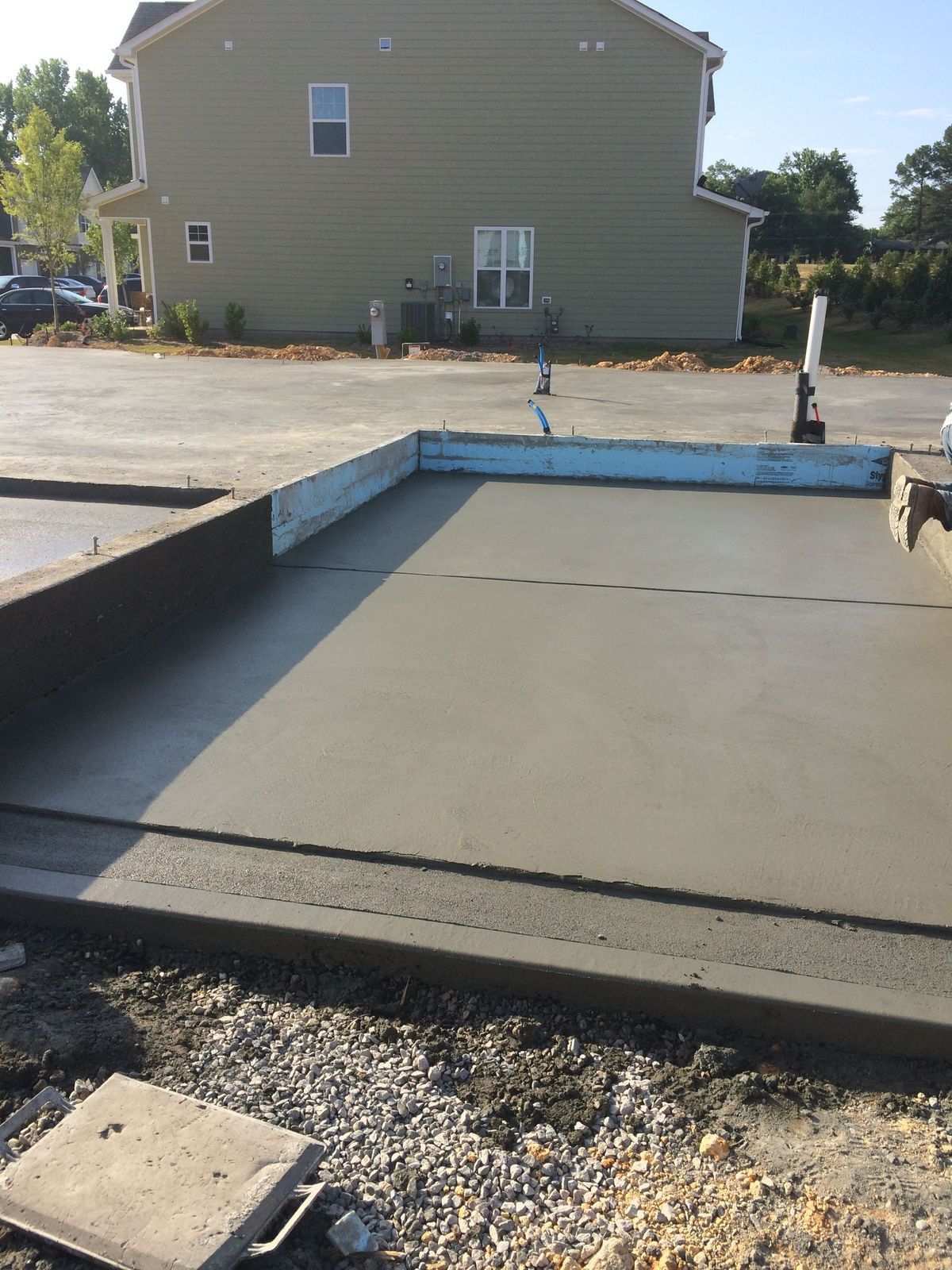 A freshly poured concrete slab foundation with blue foam insulation edges, situated in a gravel lot near a suburban home.