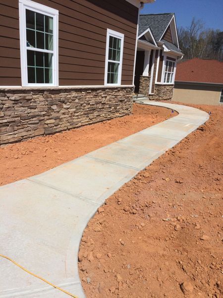 A concrete walkway leads to the entrance of a house with brown siding and stone veneer, surrounded by exposed red dirt.