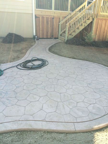 A curved, stamped concrete patio with a flagstone pattern leads to wooden deck stairs beside a house.