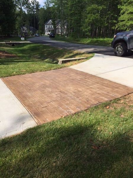 A rectangular section of brown patterned brick pavers inset into a light concrete driveway beside a grass lawn and street.