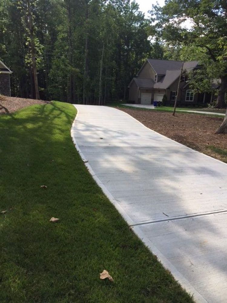 A concrete driveway curves through a residential yard with grass on the left and trees and a house in the background.