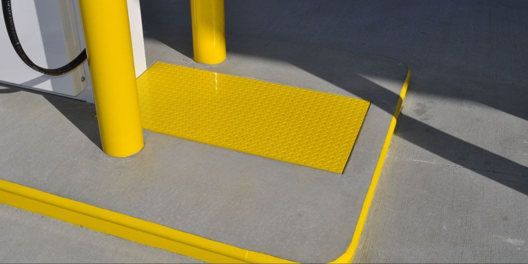 A yellow, textured tactile paving mat installed on a concrete gas station island near yellow safety bollards.
