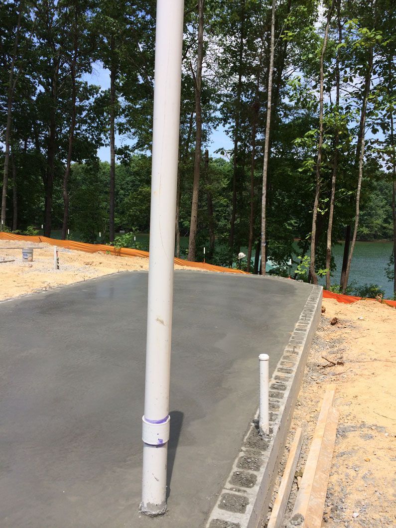 A new concrete slab foundation with plumbing pipes protruding, overlooking a wooded area and lake.