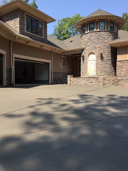 A tan house with stone accents, a turret entry, and a large, empty concrete driveway on a sunny day.
