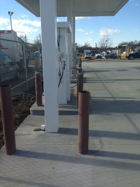Gas pump island under a canopy with metal protective bollards on a concrete lot.
