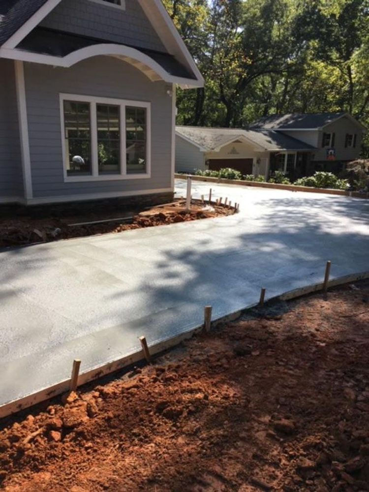 A freshly poured concrete driveway in front of a gray house with a wooden frame and exposed red soil in the foreground.