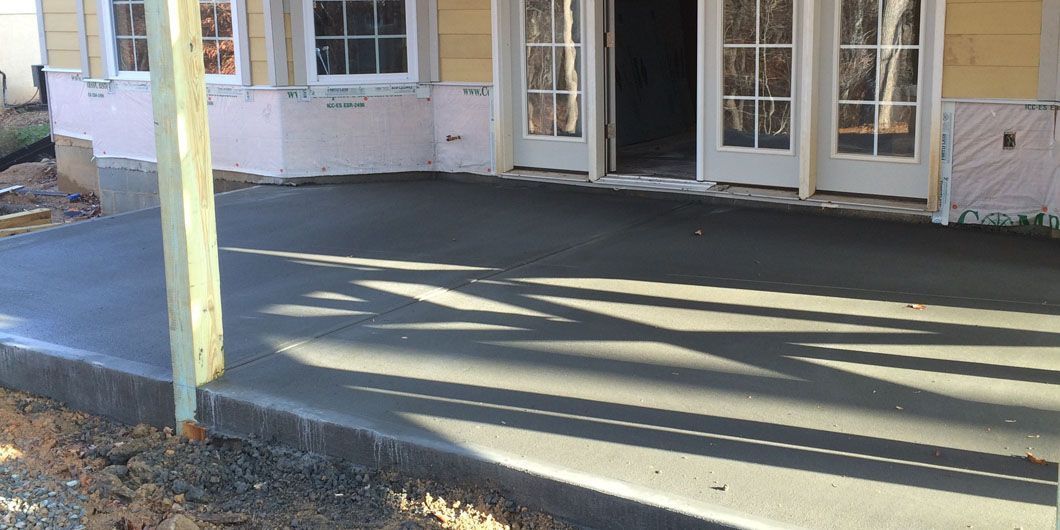 A newly poured concrete patio attached to the back of a house with a wooden support post in the foreground.
