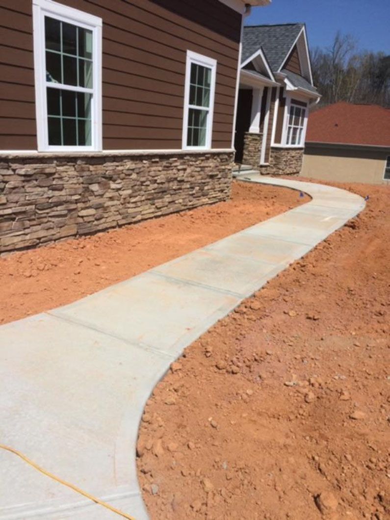 A new concrete walkway leads to the entrance of a house with brown horizontal siding and a stone-veneer base.