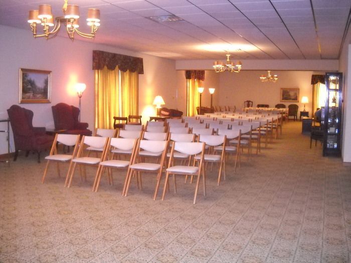 Interior view of a formal room with rows of chairs, chandeliers, and paintings, likely a funeral home.