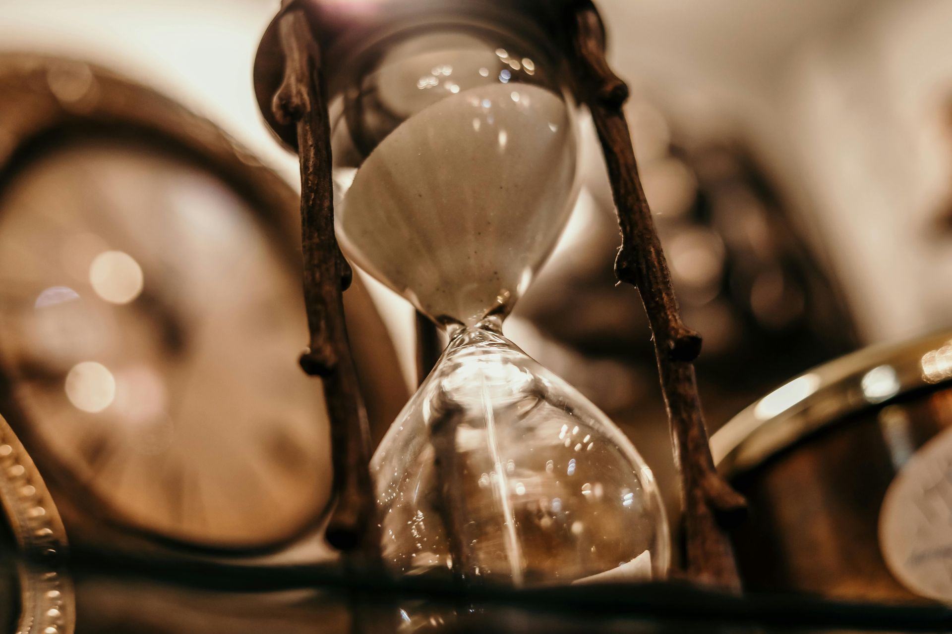 Hourglass with brown frame, sand flowing, clock face and blurred objects in background.