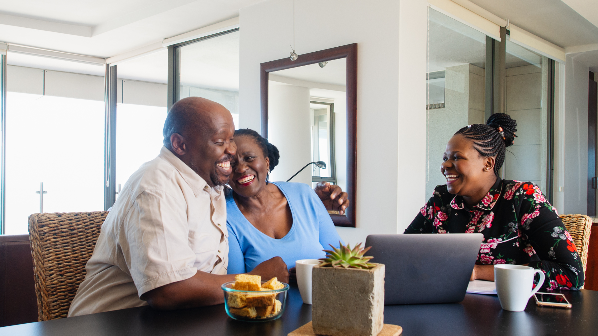 Couple smiles, arm around each other, with a woman at a table with laptop, plant and mugs.