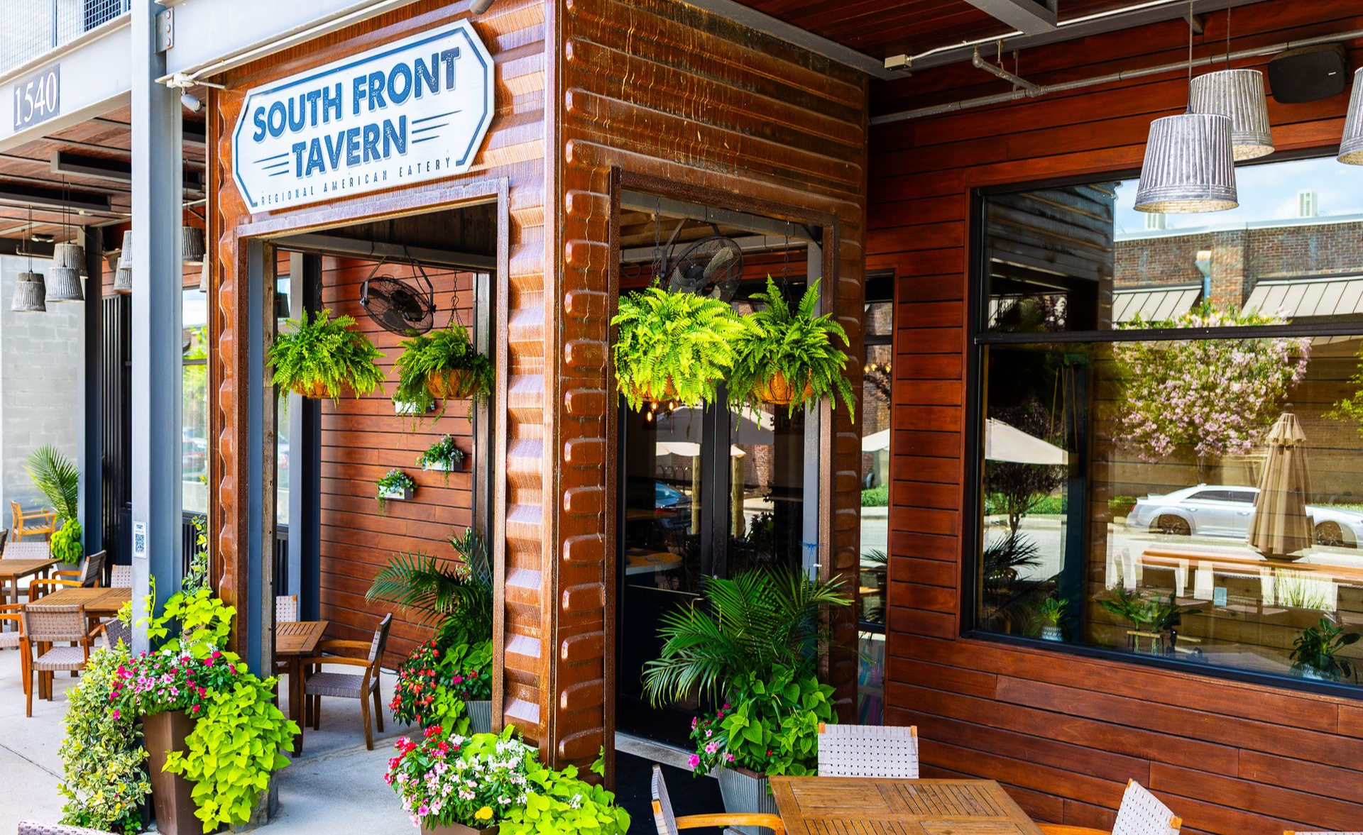 Entrance of South Front Tavern with brick facade, hanging plants, and large windows on a sunny street.