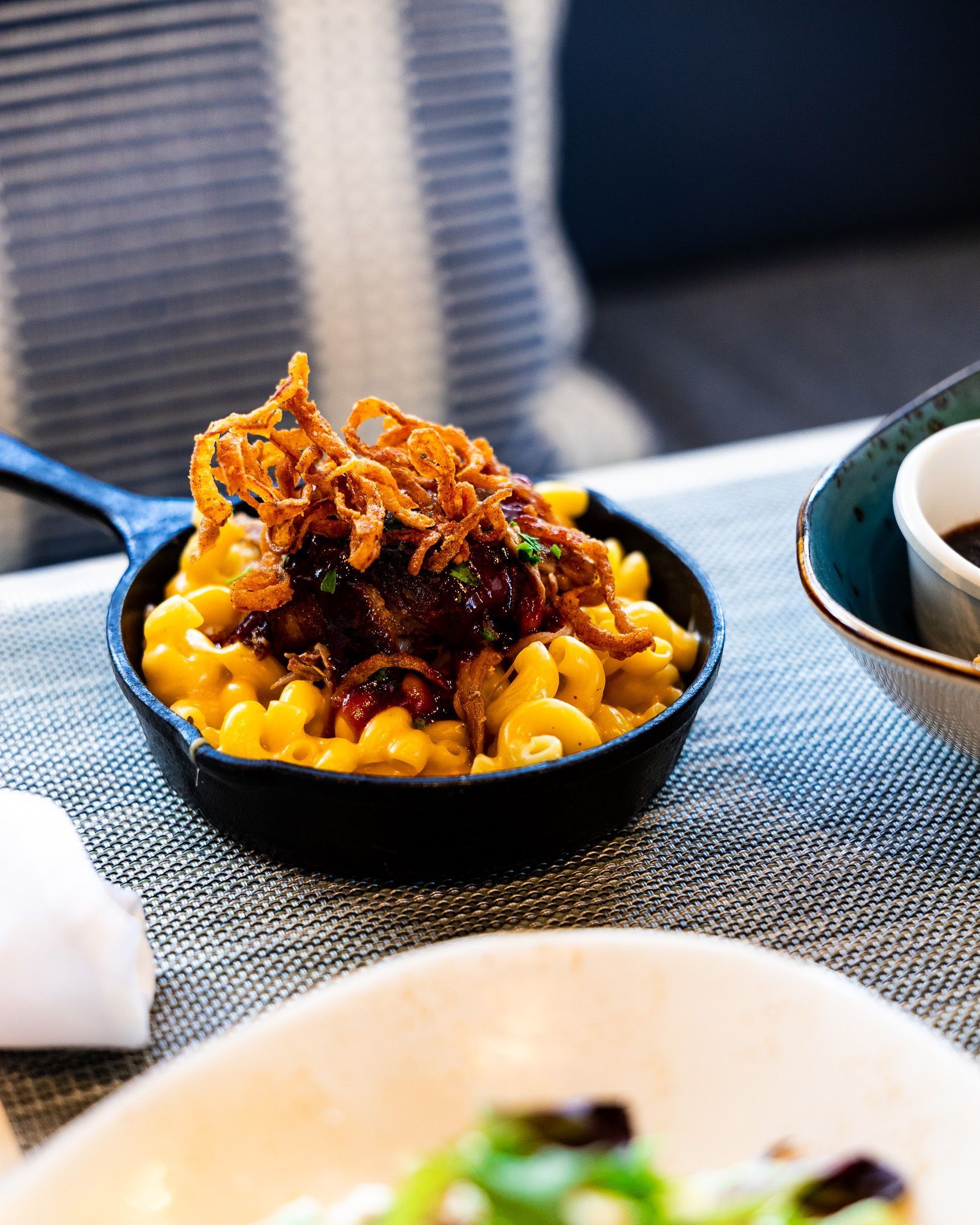 Black bowl of macaroni topped with fried garnish on a dining table beside a teacup