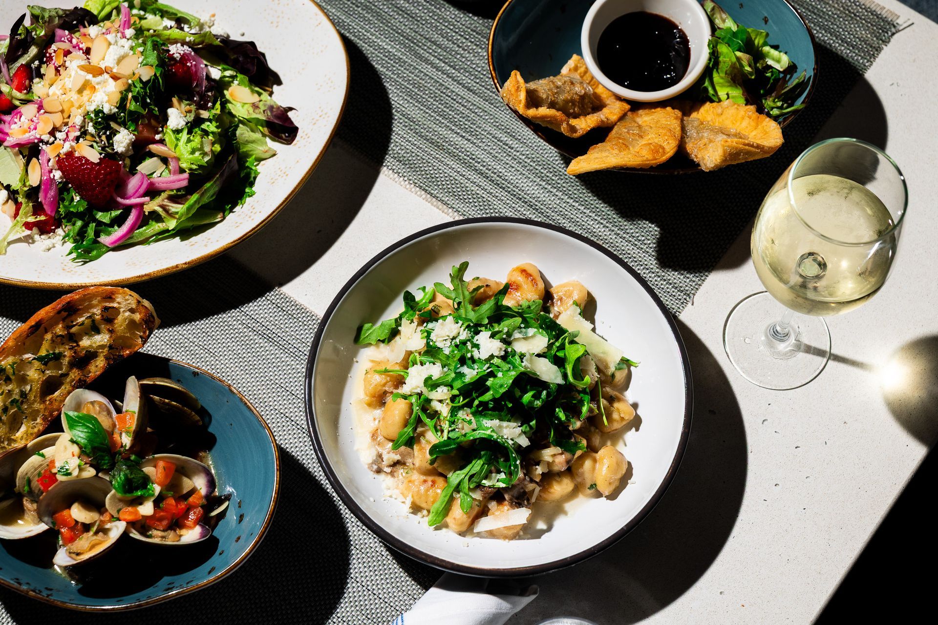 Assorted gourmet dishes and a drink on a sunlit table with dramatic shadows