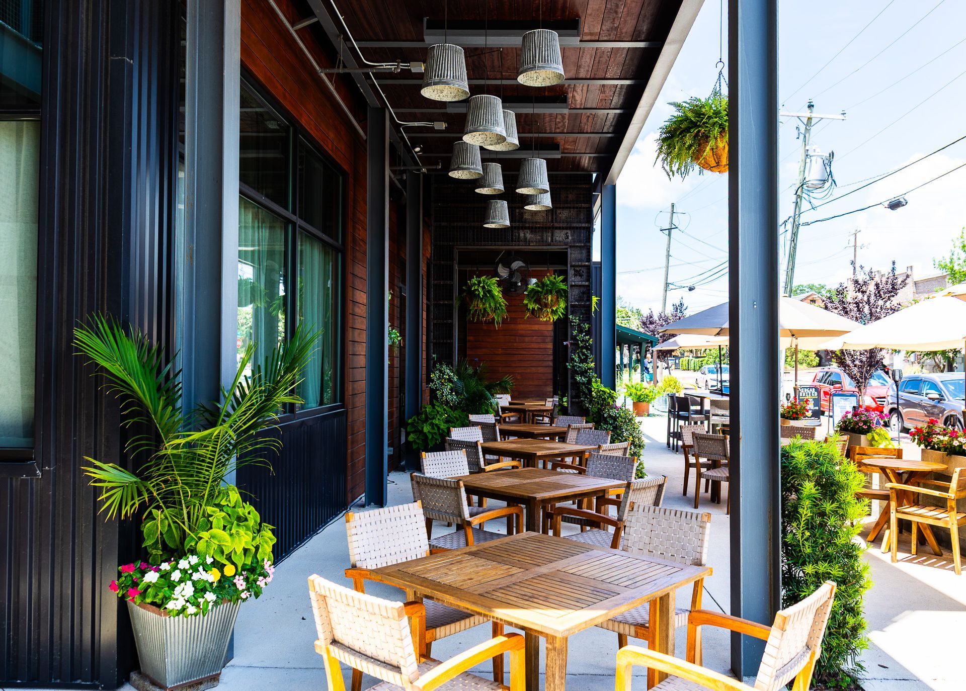 Outdoor café patio with wooden tables, potted plants, and a street view in bright daylight