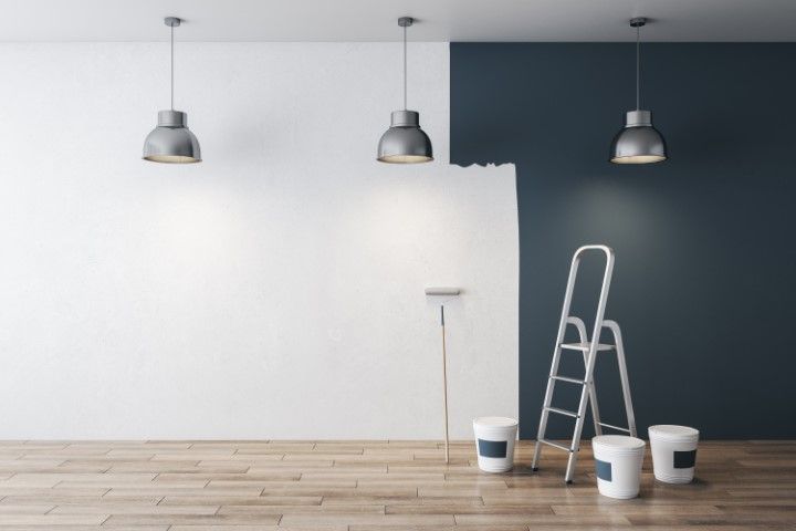 Paint roller and ladder beside a dark navy wall being painted in a room with hanging lights and wood floor