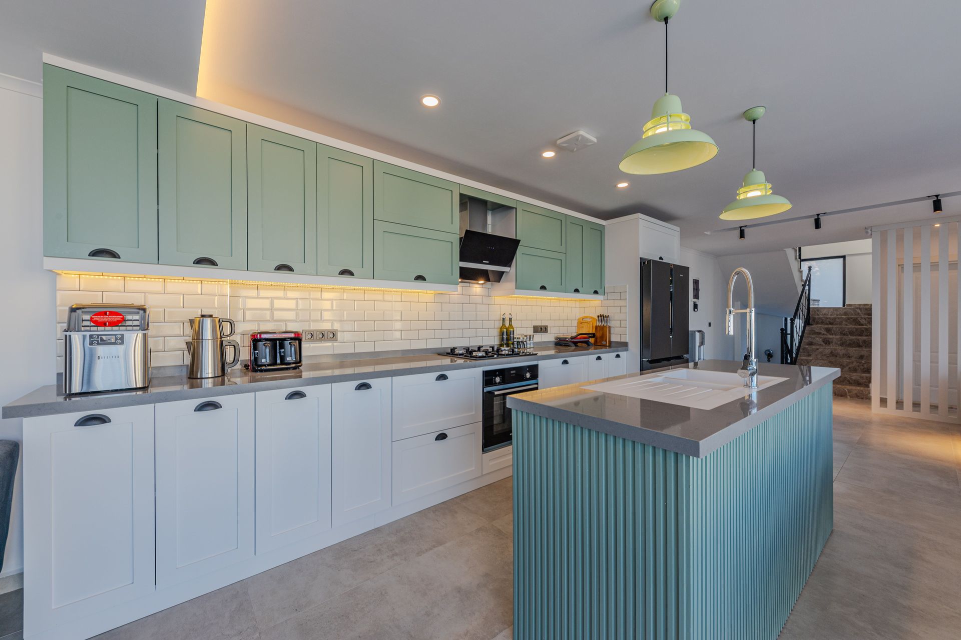 Modern kitchen with pale green cabinets, white counters, and a large teal island under pendant lights.