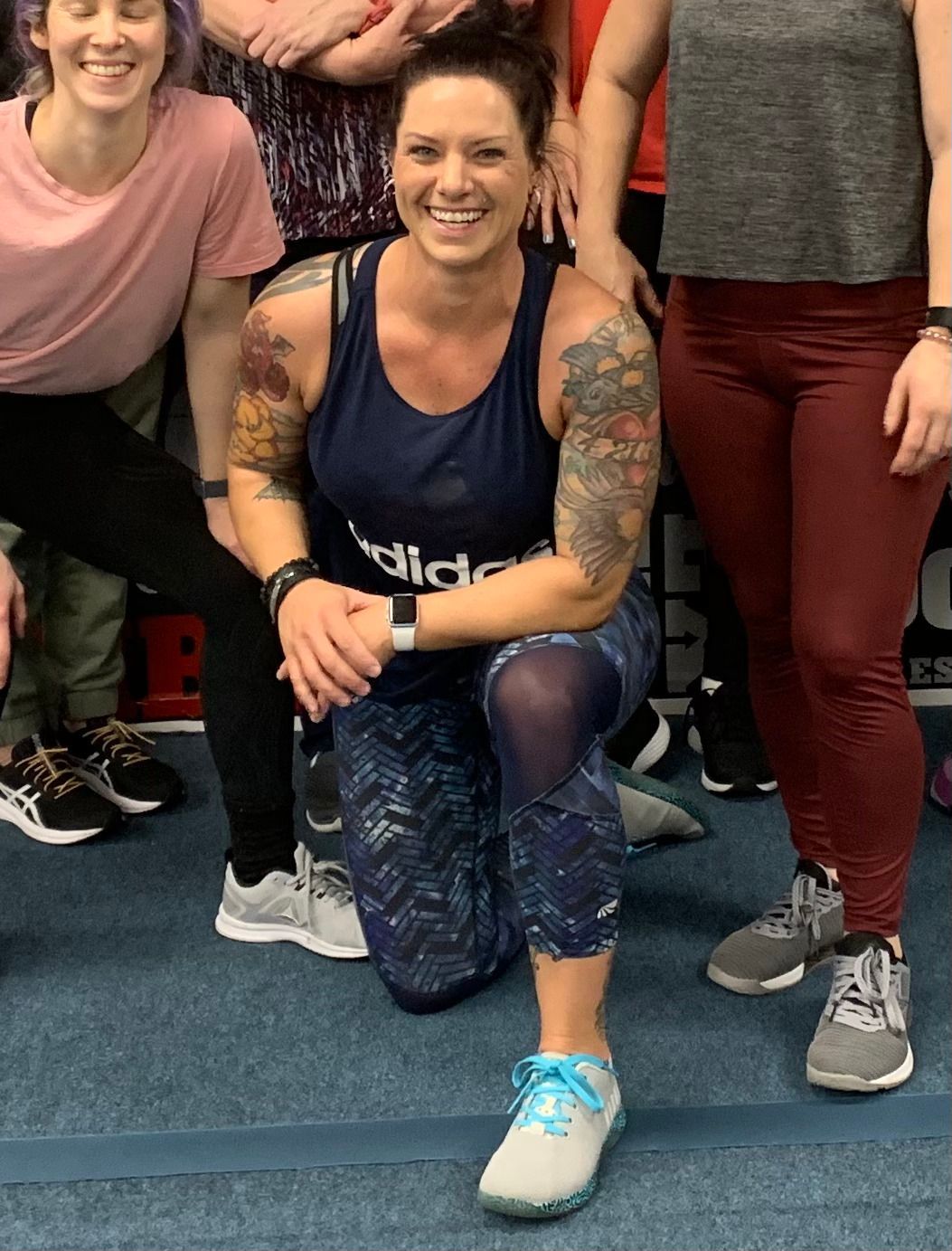 Woman with tattoos kneels, smiling, in a gym. Wearing Adidas tank, patterned leggings, and sneakers. Surrounded by other people.