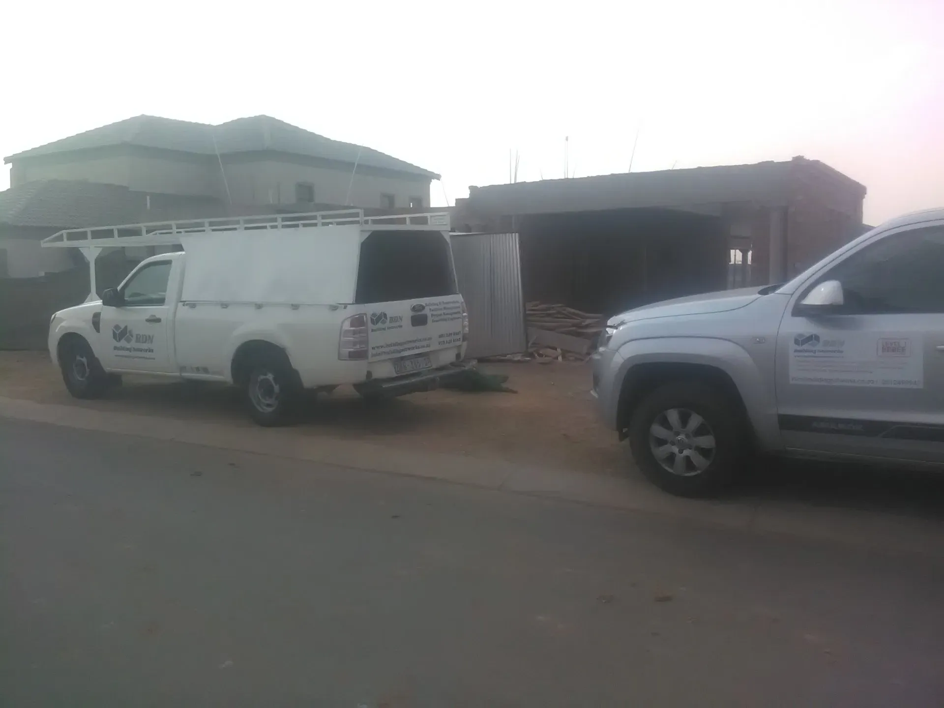 Two white trucks are parked in front of a house under construction.