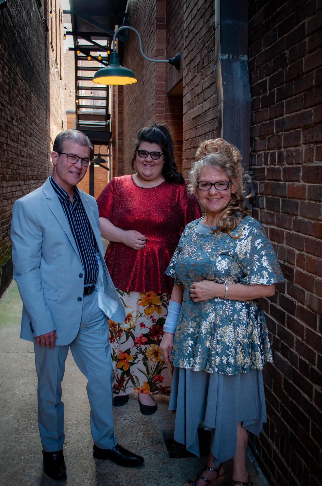 Three people posing in a narrow brick alleyway. Man in blue suit, woman in red sequin top, woman in silver top.