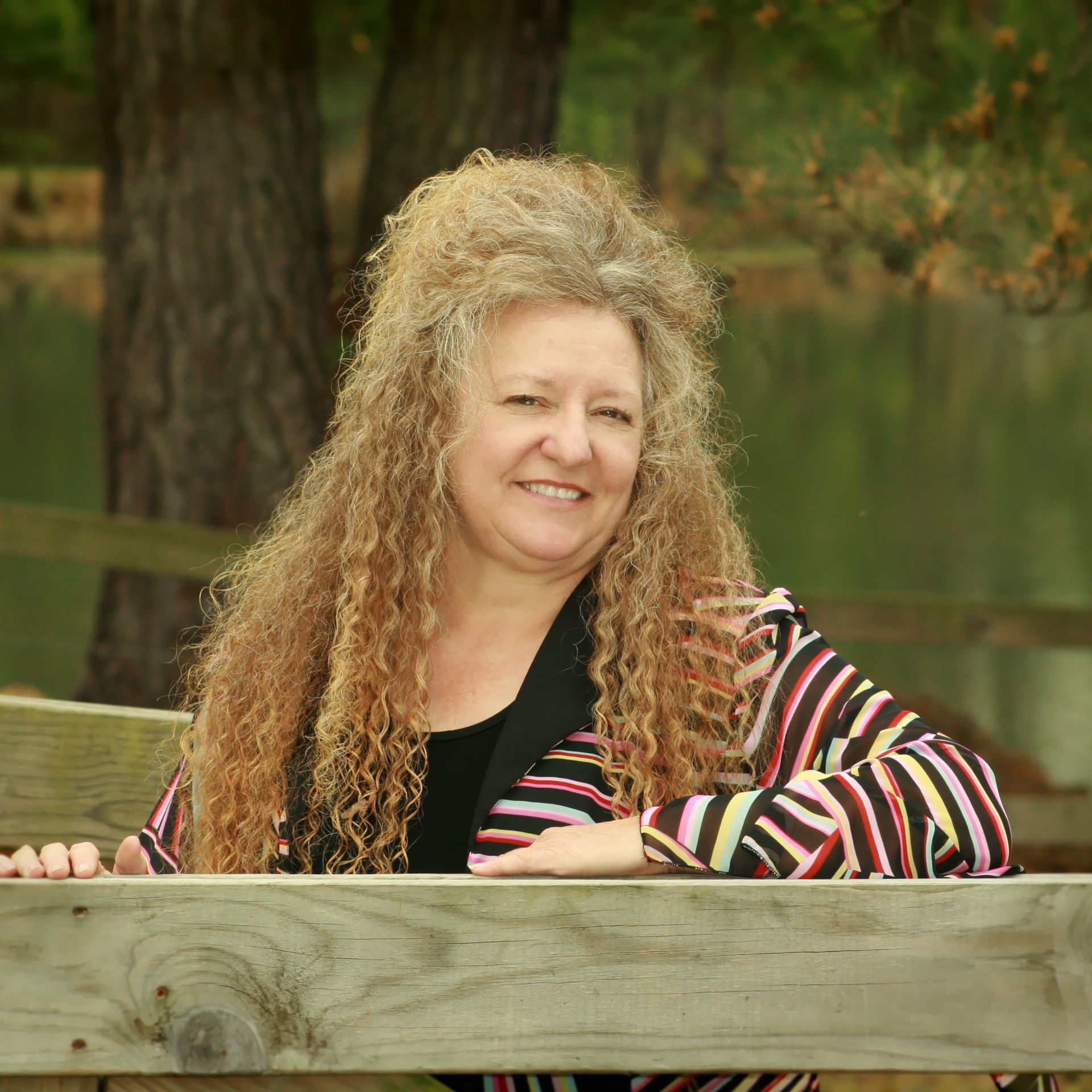 Woman with curly hair smiles, leaning on a wooden fence, wearing a striped jacket outdoors.