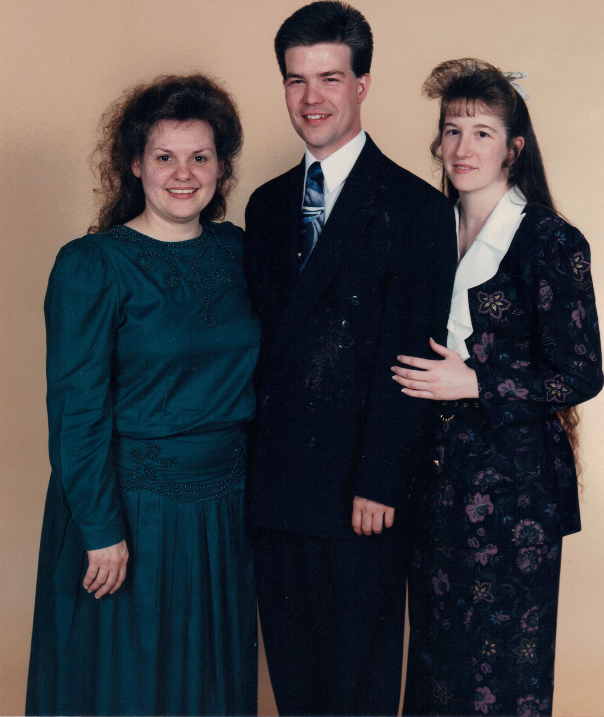 Three people standing. A man in a suit flanked by two women in dresses.