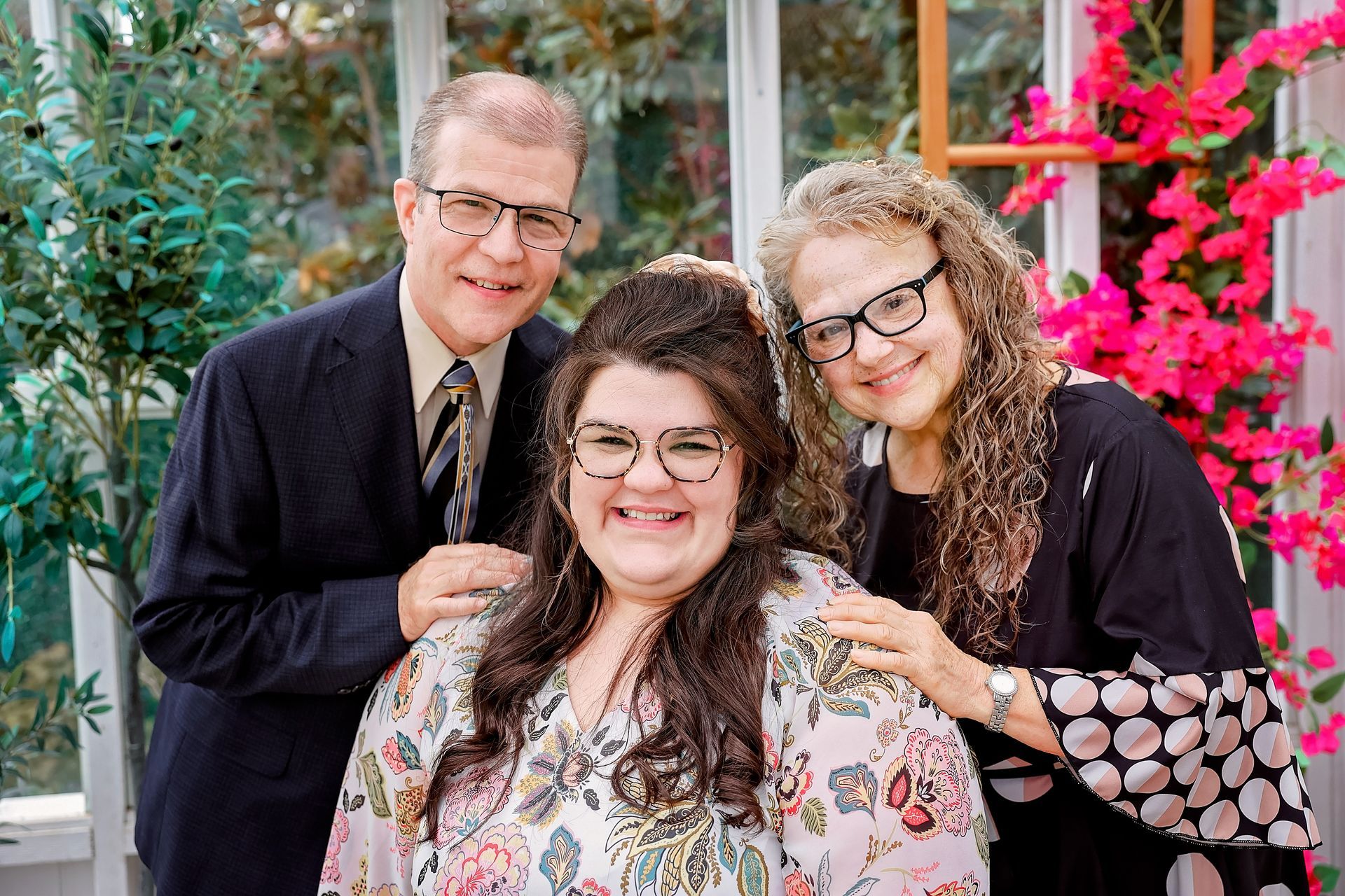 Three people smiling, posed together outdoors. Two on the sides, one in the middle, all wearing glasses.