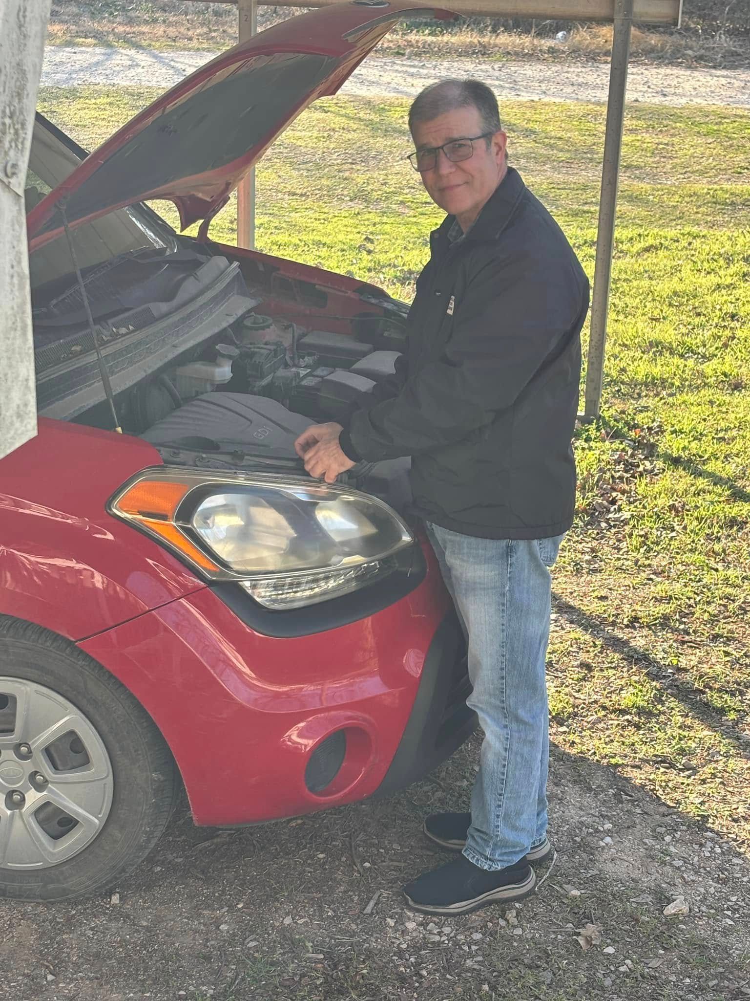 Man in glasses and jacket working on red car's engine, outdoors.
