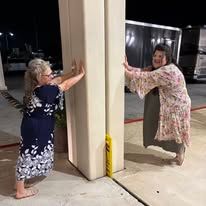 Two women push a large, beige pillar, one wearing a floral dress, the other a robe. Nighttime.