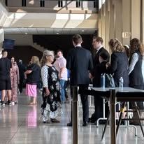 People in an atrium, some walking, some at tables. Includes a woman in a gown, suits, and glass doors.
