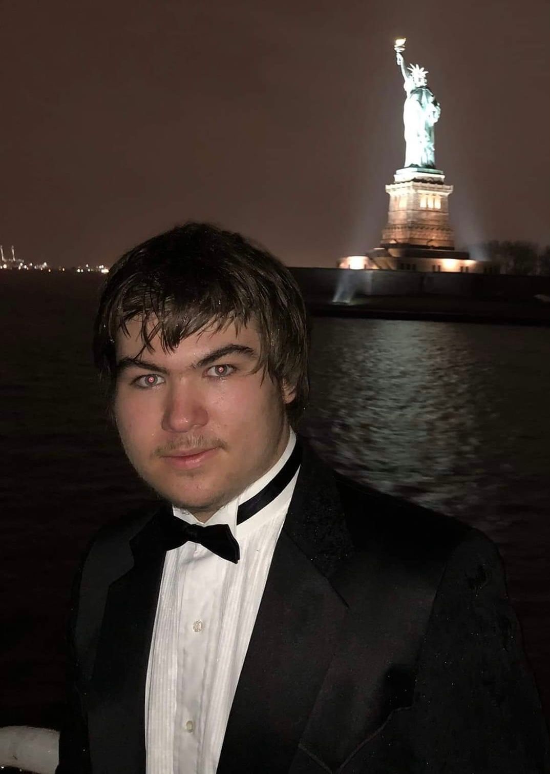 Man in tuxedo, Statue of Liberty lit up in background at night.
