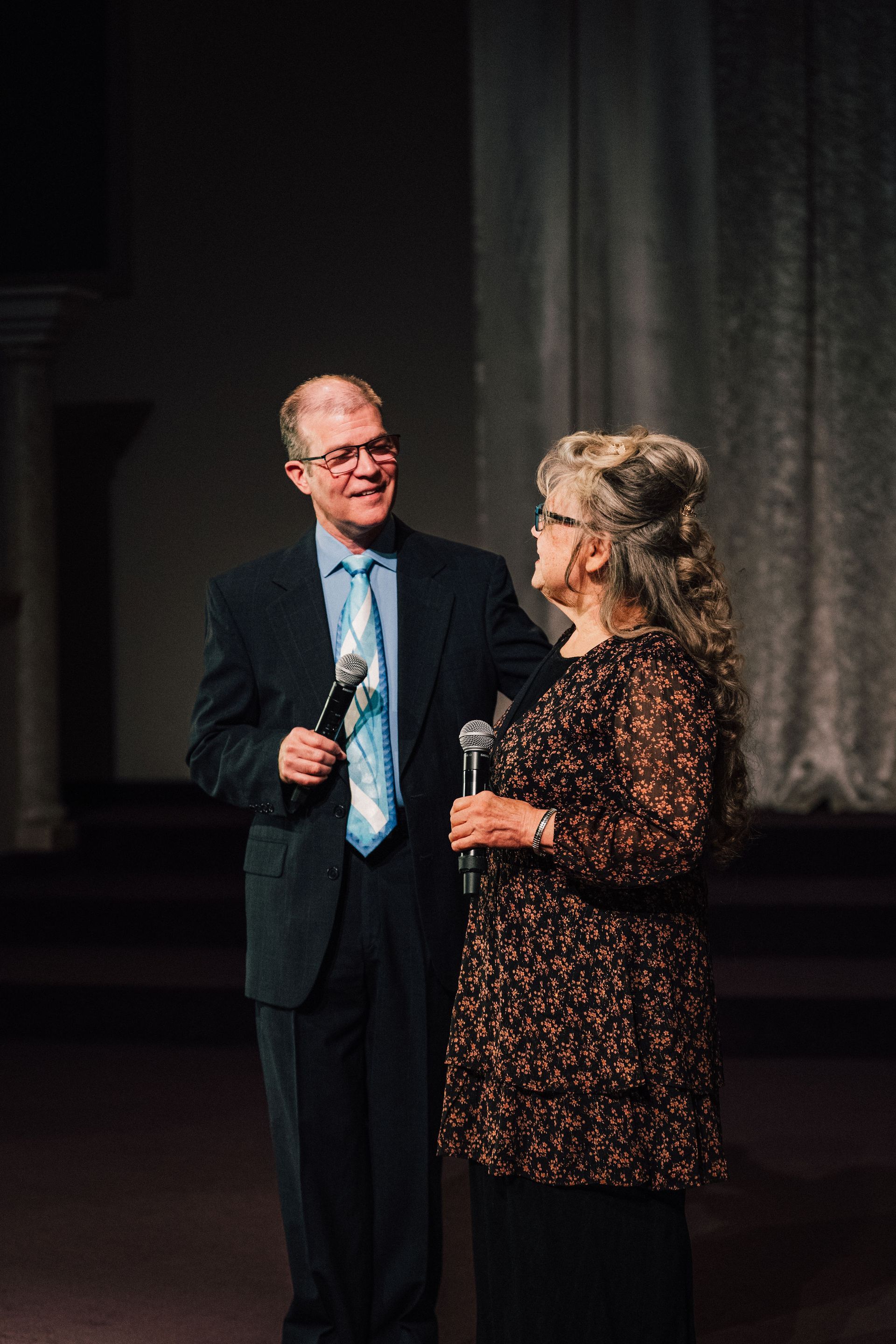Man and woman on stage, holding microphones, looking at each other, dark clothing, stage background.