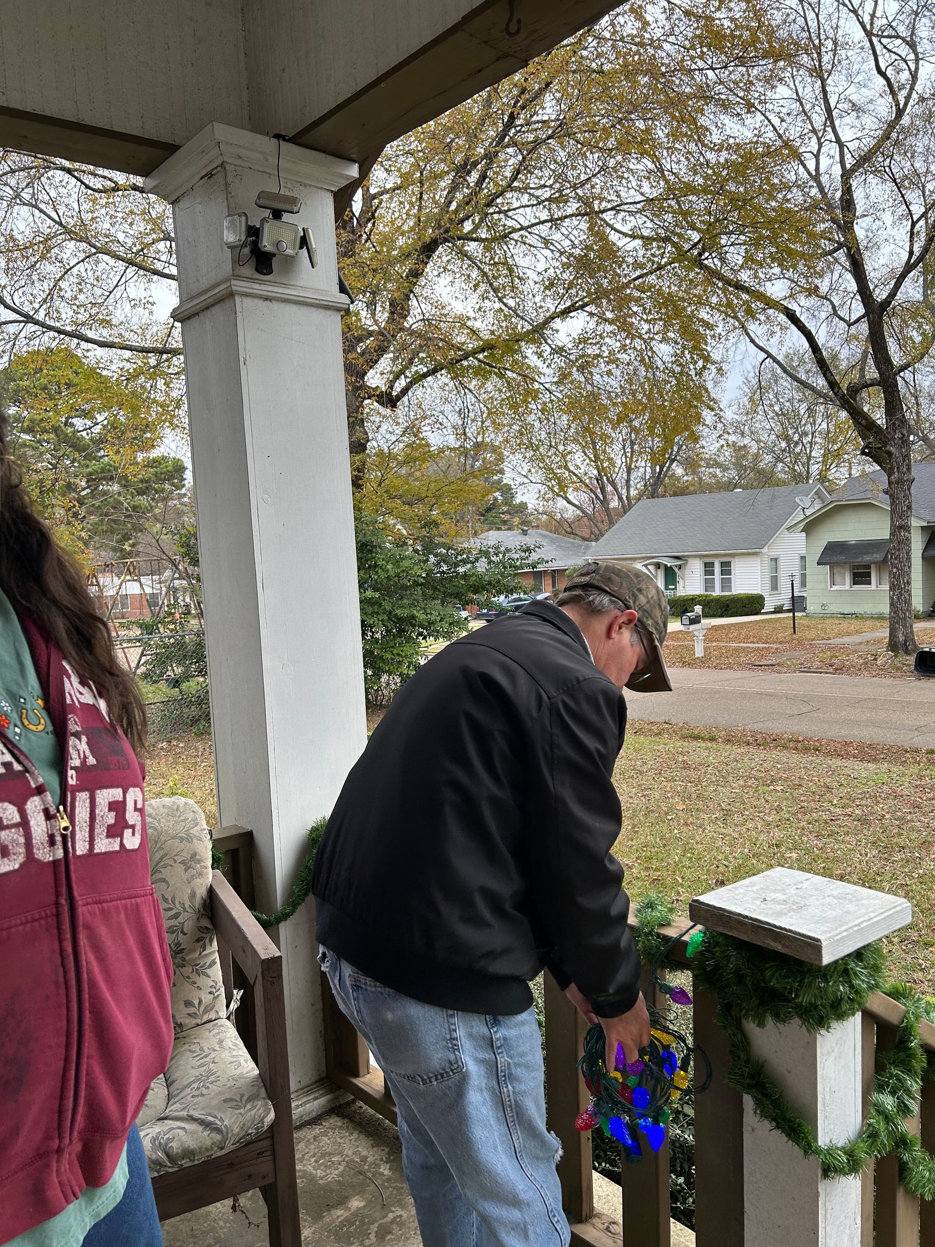 Man decorating porch with garland, autumn leaves in background.
