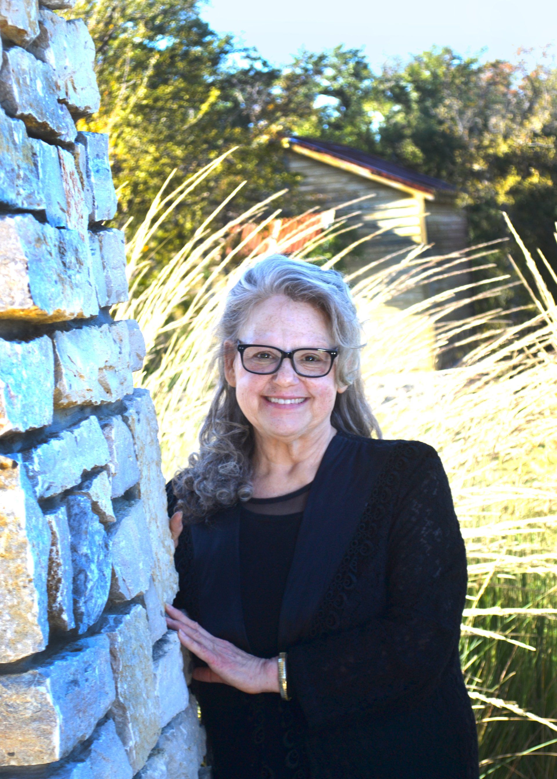 A woman wearing glasses is leaning against a stone wall.