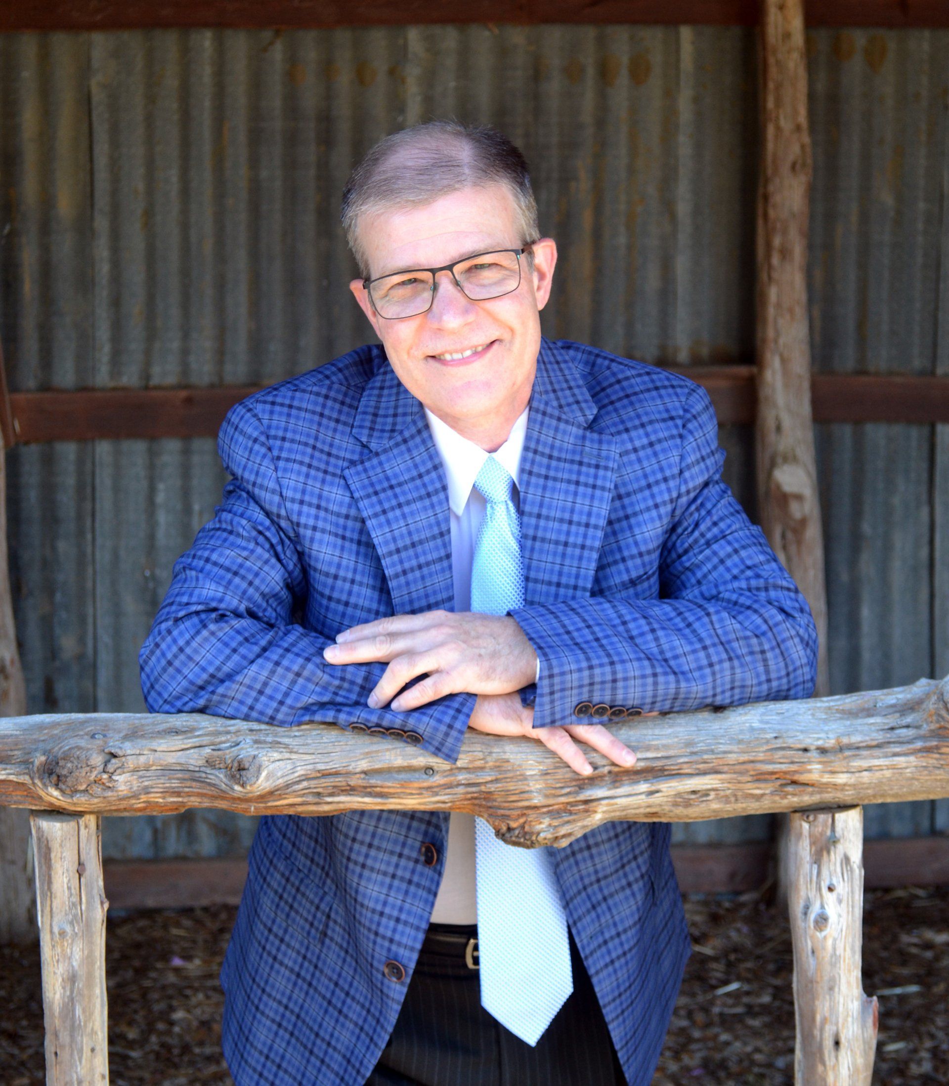 A man in a blue suit and tie is leaning on a wooden fence.