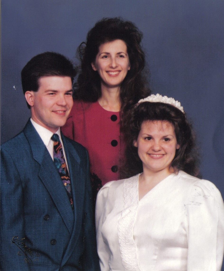 Three people in formal wear pose against a blue backdrop. A woman in a white dress smiles, while a man in a blue suit and a woman in a red top also smile.
