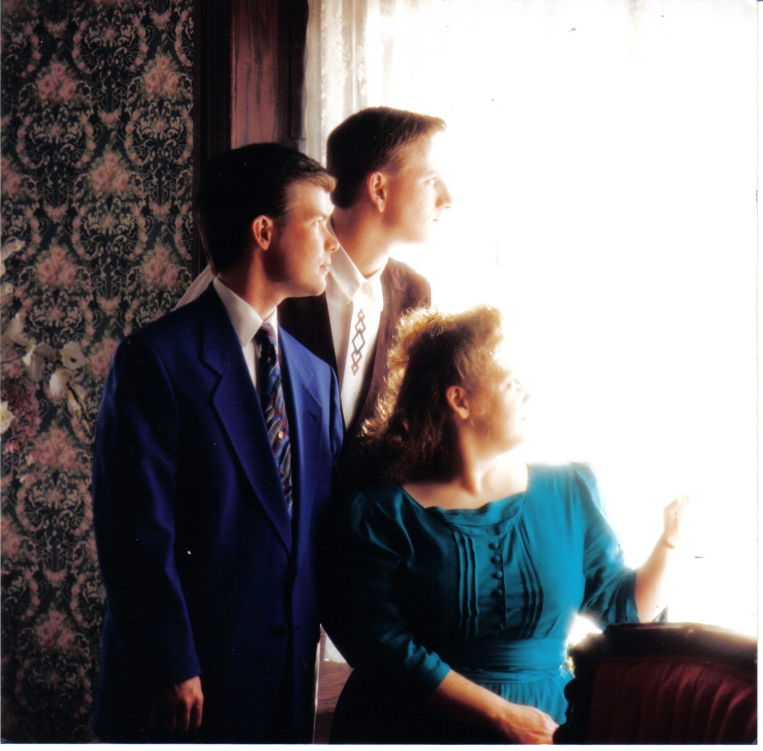 Three people looking out a bright window. A man in blue suit and tie, a man in a sweater, and a woman in a teal dress.