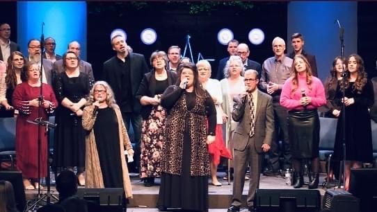 Choir singing on stage with spotlights; individuals holding microphones, some wearing formal attire.