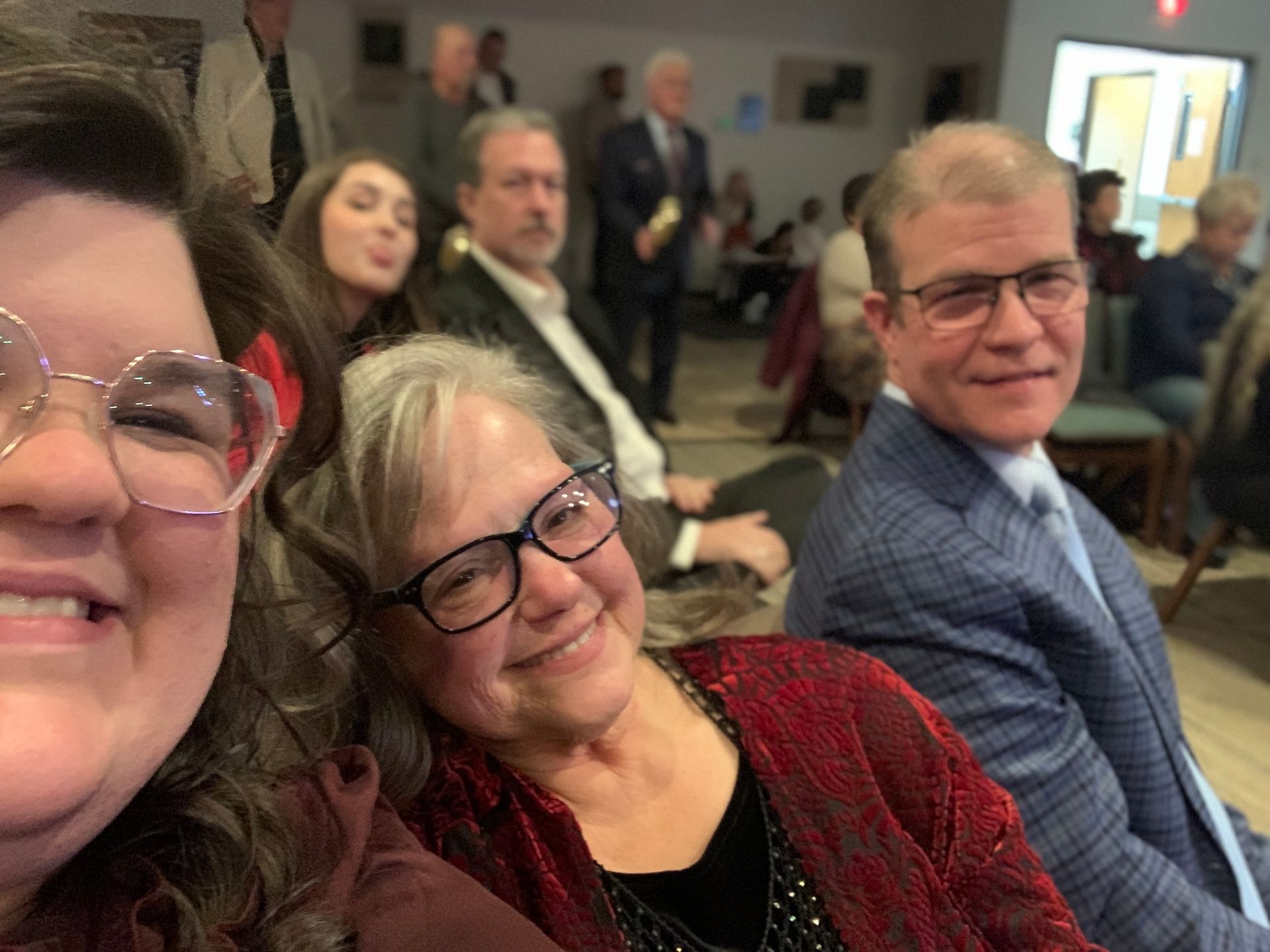 People taking a selfie in a room with a crowd. Woman with glasses and a red outfit smiles. Other people in the background.