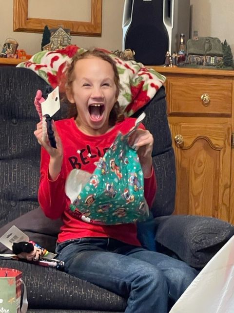 A little girl wearing a red shirt that says reindeer is sitting in a chair