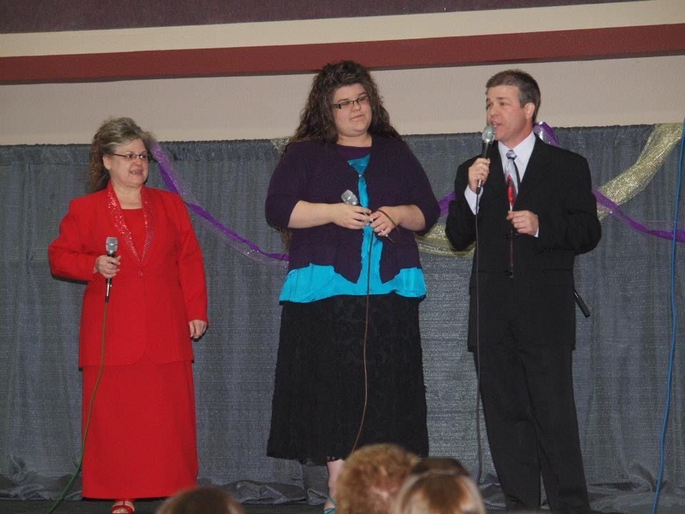Three people on stage with microphones. Woman in red suit, woman in blue and purple, man in suit.