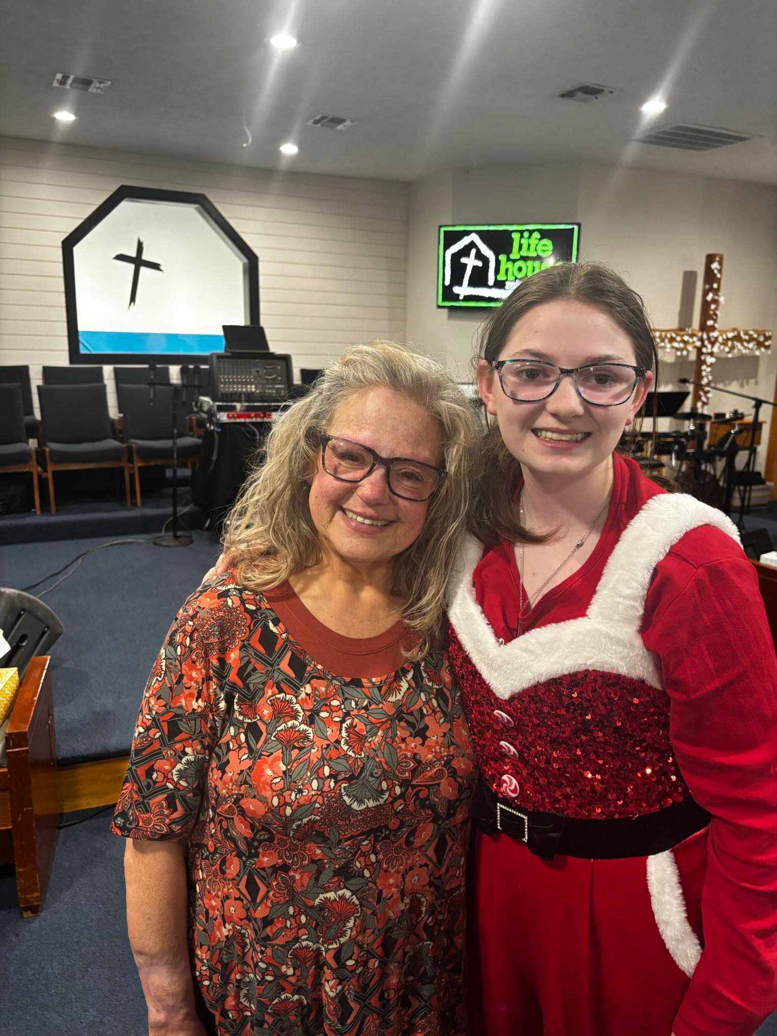 older lady hugging younger in front of a church cross