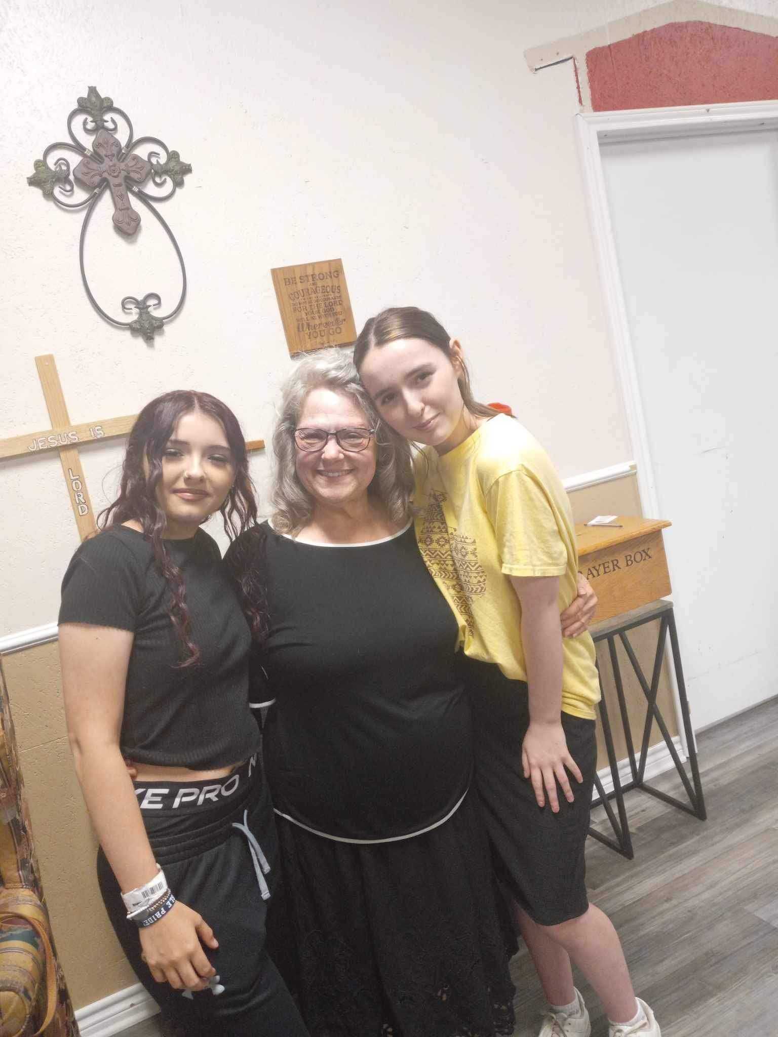 Three women are posing for a picture in a room with a cross on the wall.