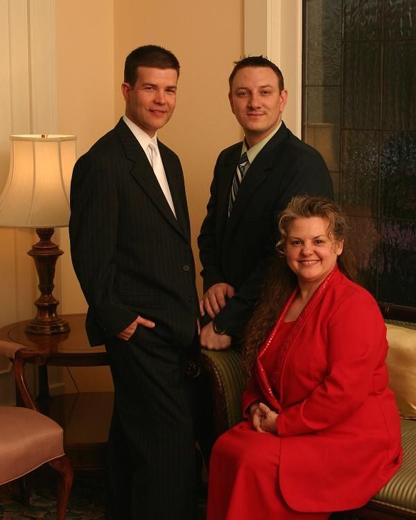 Three people posed formally indoors: two men in suits and a woman in a red dress.