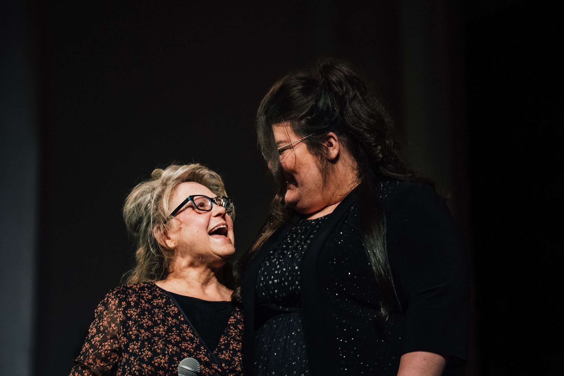 Two women on stage, smiling and laughing. Dark clothing, one with glasses, dark background.